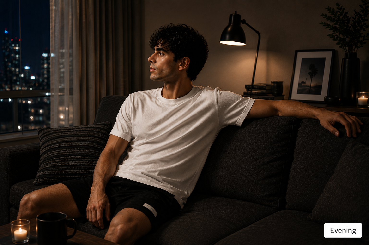 A man wearing a white crewneck t-shirt and black athletic shorts sits on a dark couch in a dimly lit living room at night, looking out a window with a city skyline visible.